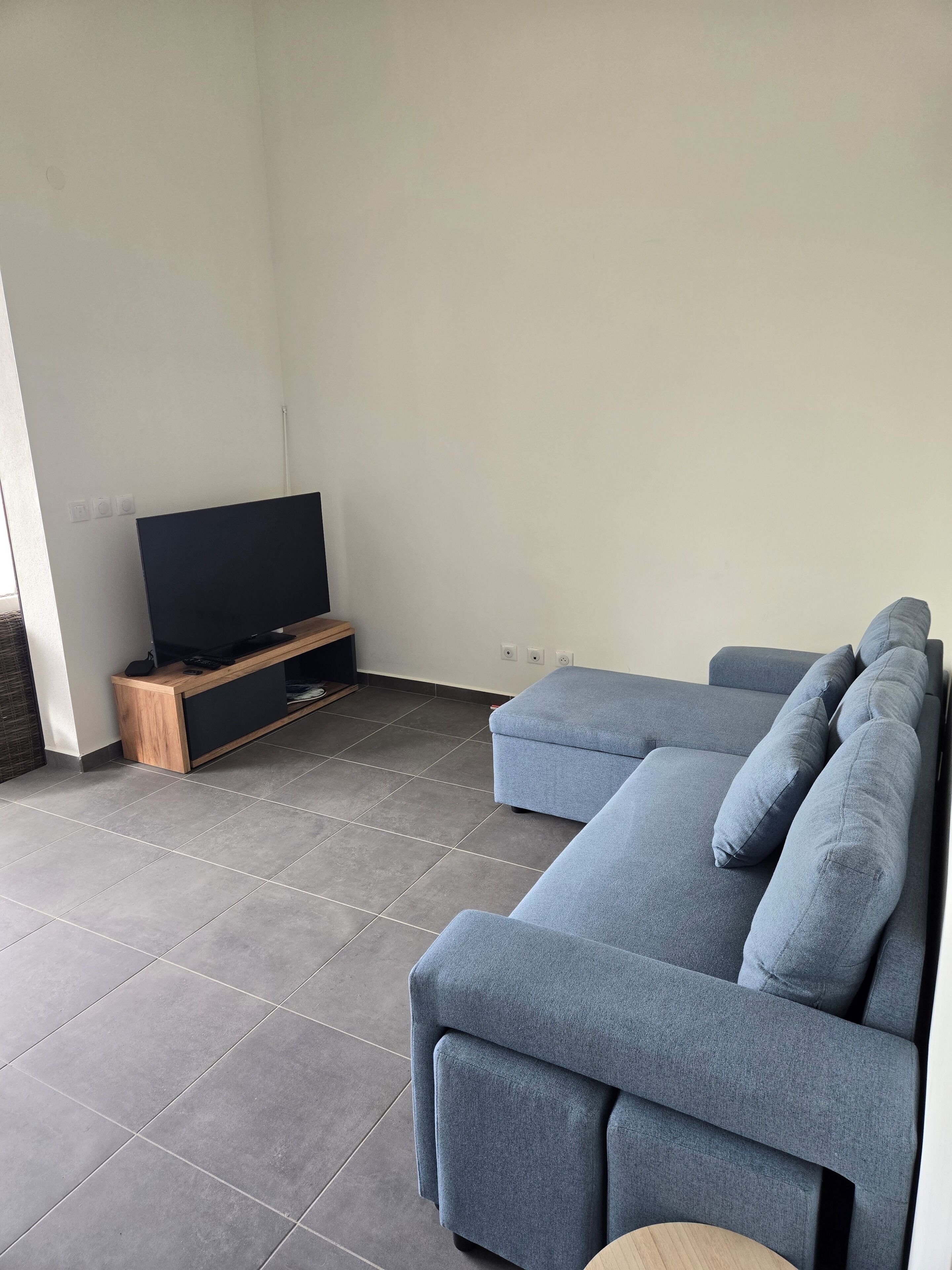 Una sala de estar moderna con un sofá seccional, un televisor de pantalla plana sobre un soporte de madera y piso de baldosas.