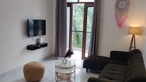 Uma sala de estar moderna com um ventilador de teto, uma TV de tela plana, um sofá escuro, um banquinho redondo e uma mesa de centro de vidro.