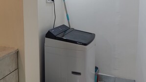 Quarto compacto com parede branca, uma lâmpada no teto, um cesto de lixo pequeno e um eletrodoméstico compacto.