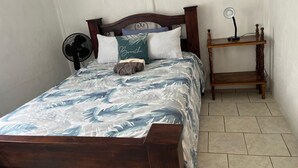 Un letto con una coperta a fiori blu e bianchi, una testiera in legno, un comodino con una lampada e una ventola.