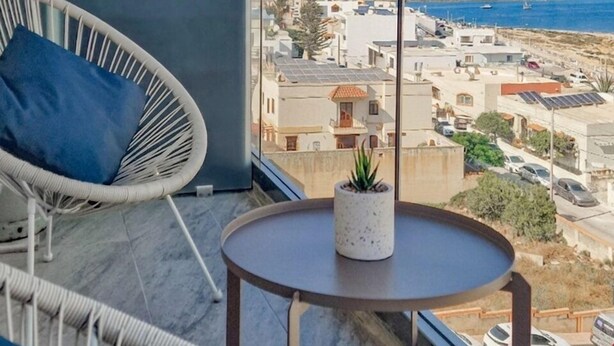Un balcón con vistas al mar, una silla blanca, una mesita con una planta y un paisaje urbano al fondo.