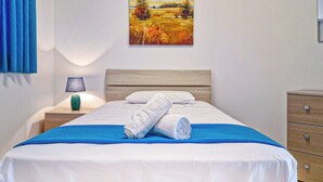 Una cama bien hecha con sábanas blancas y una manta azul, una mesita de noche de madera con una lámpara, un aparador de madera y un cuadro de un paisaje en la pared.