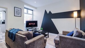 A modern living room with a flat-screen TV, two sofas, a coffee table, and a wall-mounted lamp. - Neat 4BR House | Pool Table | Free Parking (Greater Manchester)