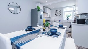 A modern kitchen with white cabinets, a black clock, and a dining area with blue tableware. - Neat 4BR House | Pool Table | Free Parking (Greater Manchester)