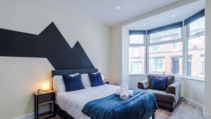 A modern hotel room with a large bed, a sofa, a nightstand, and a window with a view of buildings. - Neat 4BR House | Pool Table | Free Parking (Greater Manchester)