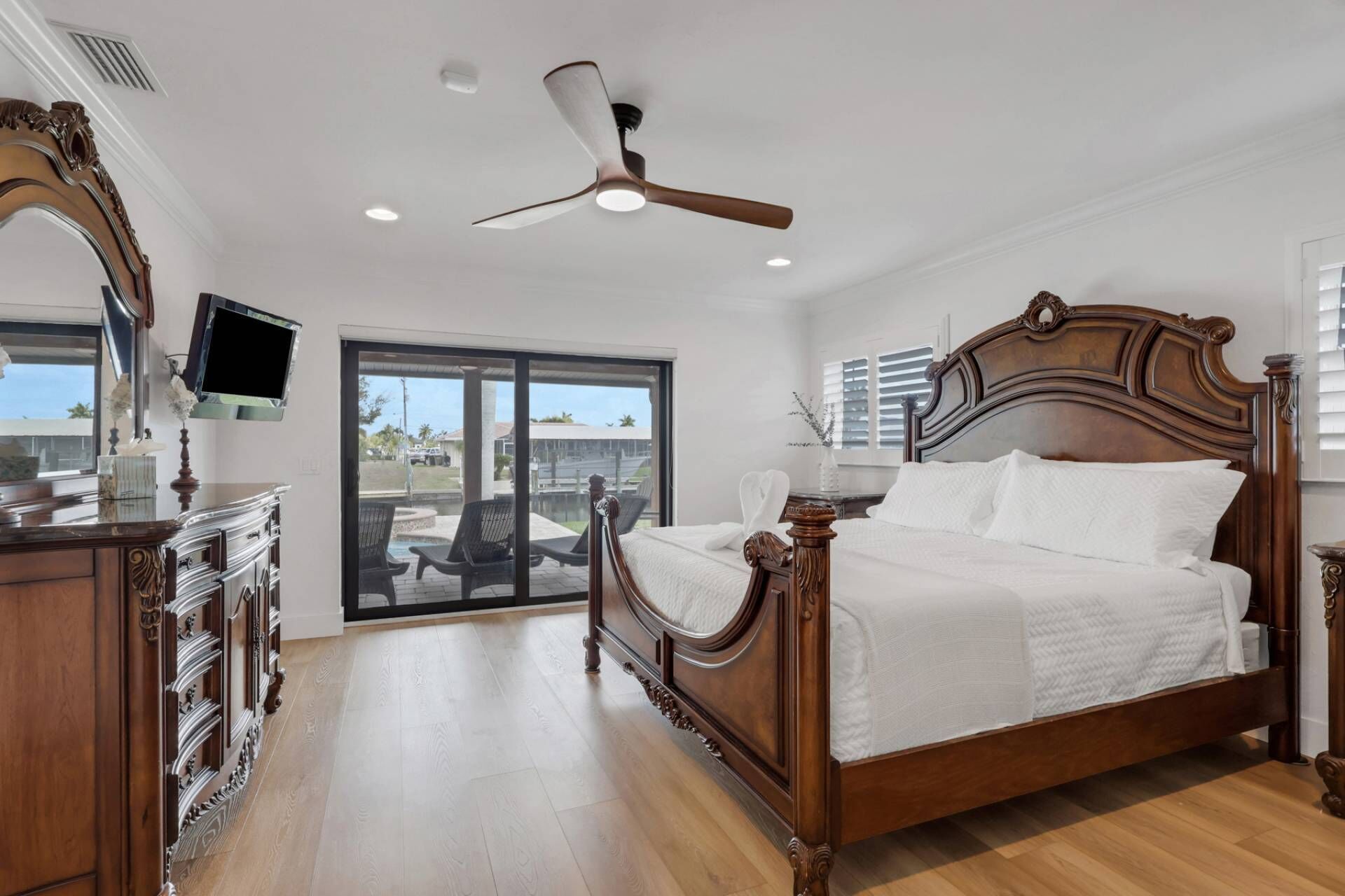 A bedroom with a wooden bed, a ceiling fan, a TV, and a view of the outdoors.