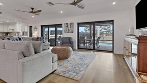 A spacious living area with a grey sofa, a woven ottoman, a patterned rug, and a dining set. There are ceiling fans, a kitchen in the background, and a pool visible through the glass doors. - Boater’s Paradise in SW Cape Coral | Coastal Retreat with Pool, Spa, Dock & Tropical Outdoor Oasis (Cape Coral)