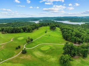 Vista aérea de un campo de golf con un lago y colinas boscosas al fondo.