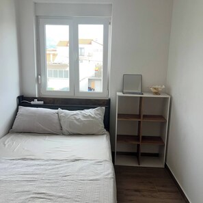 Ein Schlafzimmer mit einem Bett, einem Fenster, einem Nachttisch mit Lampe und einem kleinen Regal mit einer Dekoration.