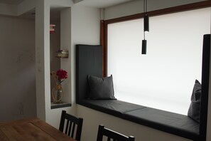 A room with a wooden table, a bench with cushions, a shelf with books and a vase with flowers, and a hanging light fixture.