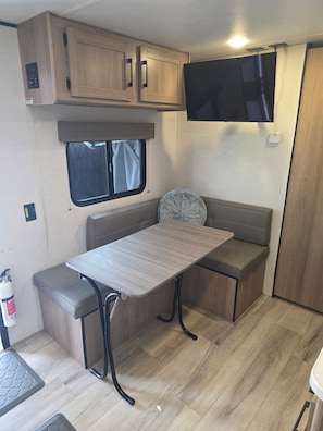 A compact RV interior with a kitchenette, a table with a bench, a window, and a mounted TV.