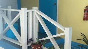 Un porche con baranda blanca, una puerta azul con la letra 'C' y plantas en macetas.