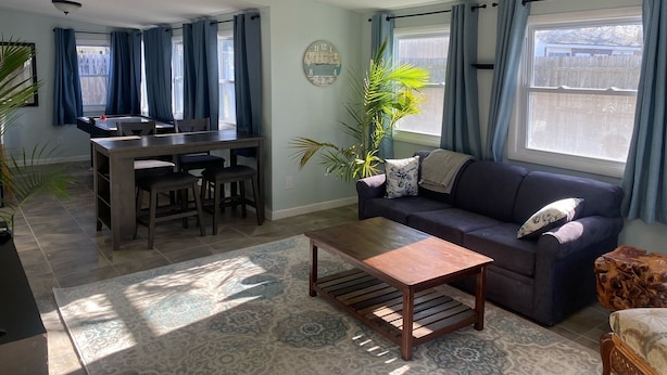 Ein Wohnzimmer mit einer Couch, einem Couchtisch, einem Esstisch, Stühlen und einem großen Fenster mit Vorhängen.