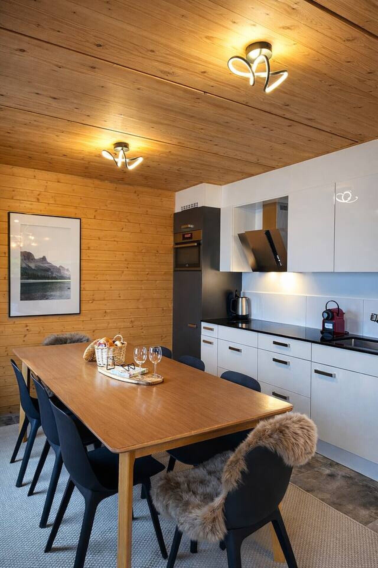 A modern kitchen with white cabinets, a wooden dining table, and a black chair with a fur throw.