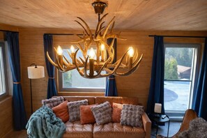 A cozy room with a leather sofa, a chandelier with antler-like design, and a view of the outdoors through a window. - Modern SPA Chalet near Interlaken • AC • Hot Tub by SwissDreamLiving (Beatenberg)