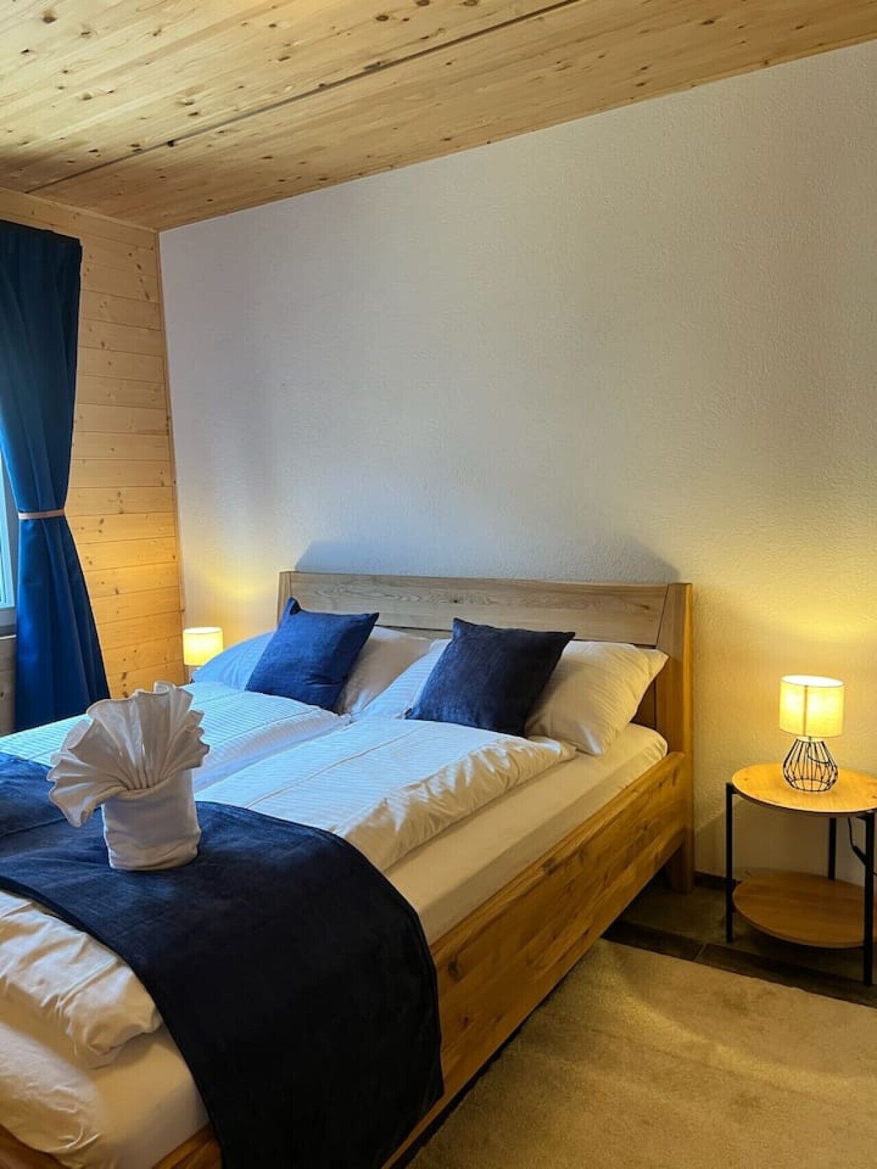 Un dormitorio con cabecera de madera, cama con ropa de cama blanca y azul, mesita de noche con lámpara y cortina azul.