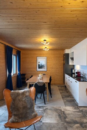 A cozy cabin interior with a wooden ceiling, a dining area with a table and chairs, a kitchen with modern appliances, and a window with curtains. - Modern SPA Chalet near Interlaken • AC • Hot Tub by SwissDreamLiving (Beatenberg)
