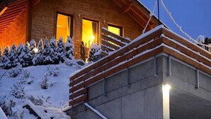 Een houten chalet met een overdekte trap naar een verlichte ingang, omgeven door een met sneeuw bedekte grond en bomen.