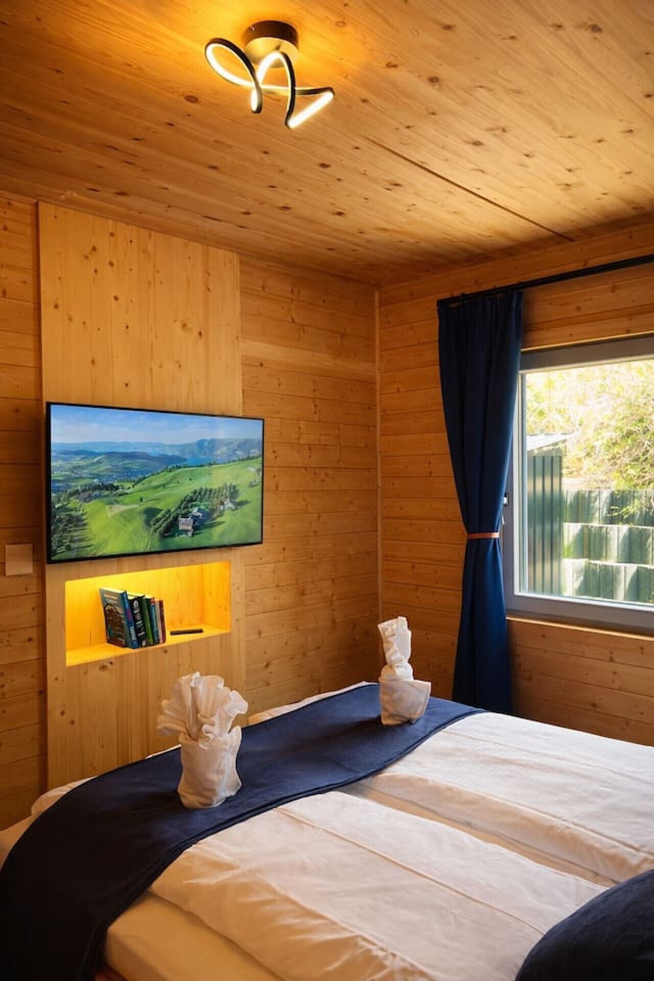 Une pièce aux murs lambrés, avec un lit, un téléviseur fixé au mur et une fenêtre avec des rideaux.