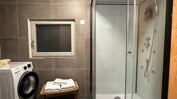 Um banheiro compacto com máquina de lavar roupa, um banquinho pequeno com toalhas e um box de vidro.