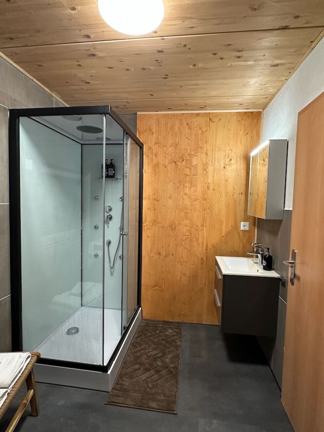 A modern bathroom with a glass shower enclosure, a wooden wall, a sink with a mirror, and a bench.