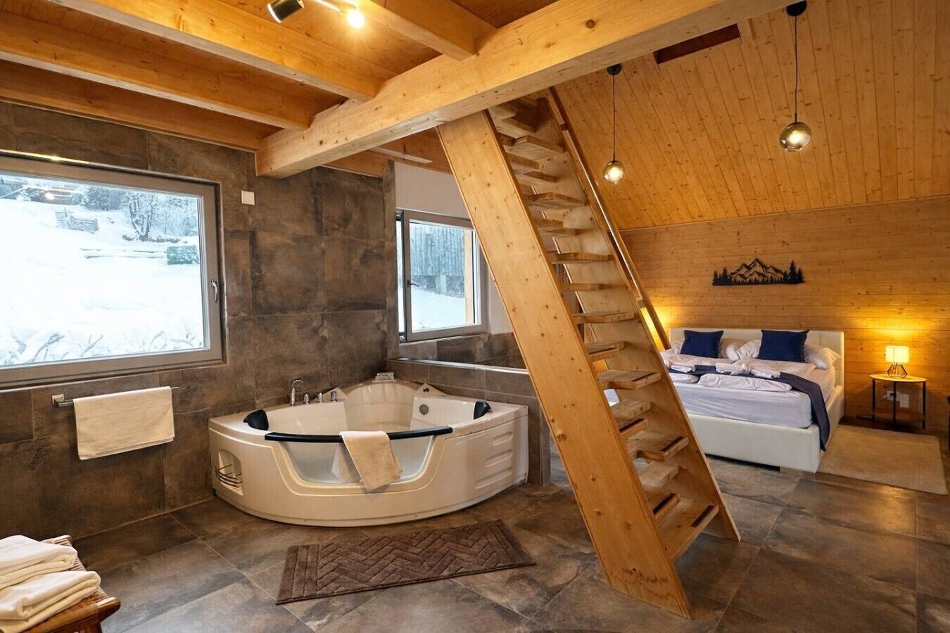 Ein modernes Interieur mit einer runden Whirlwanne, einer Holztreppe, einem Bett mit Bergdekor und Blick auf eine verschneite Landschaft durch ein Fenster.