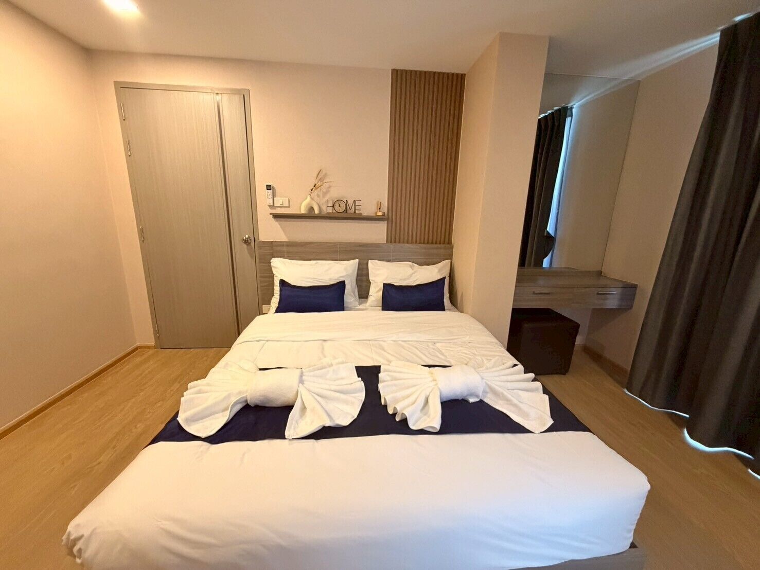 A hotel room with a bed, a door, a mirror, a shelf with a 'HOME' sign, and a window with curtains.