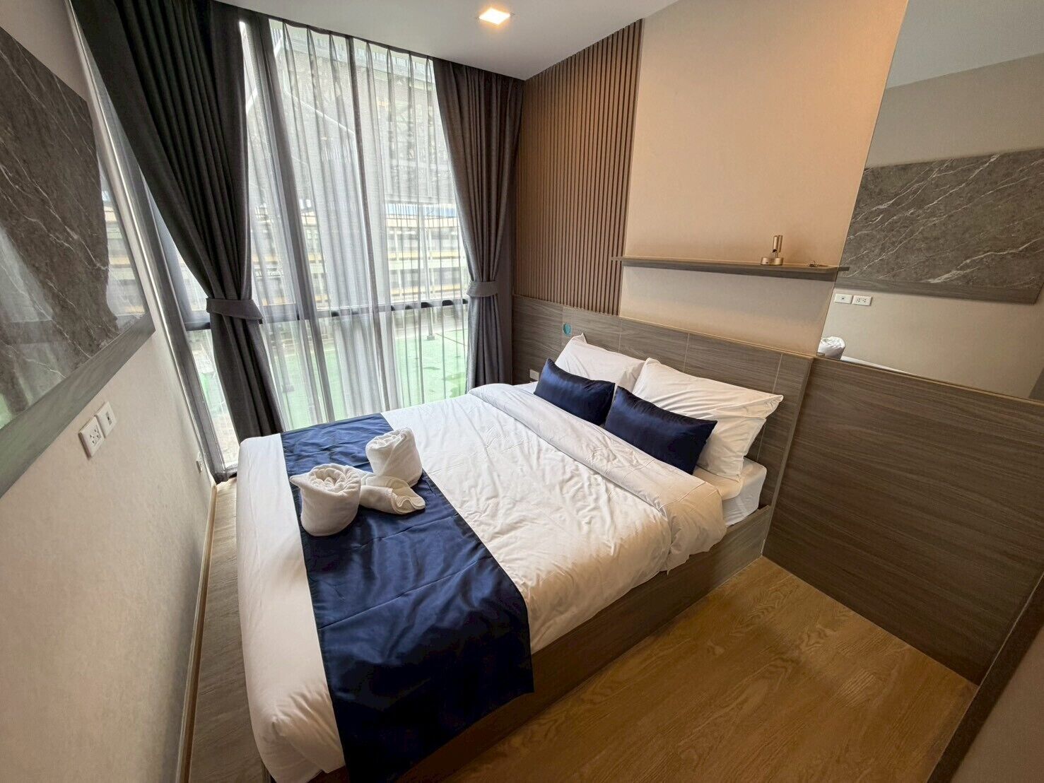 A modern hotel room with a large bed, a view of the city through the window, and a wooden headboard.