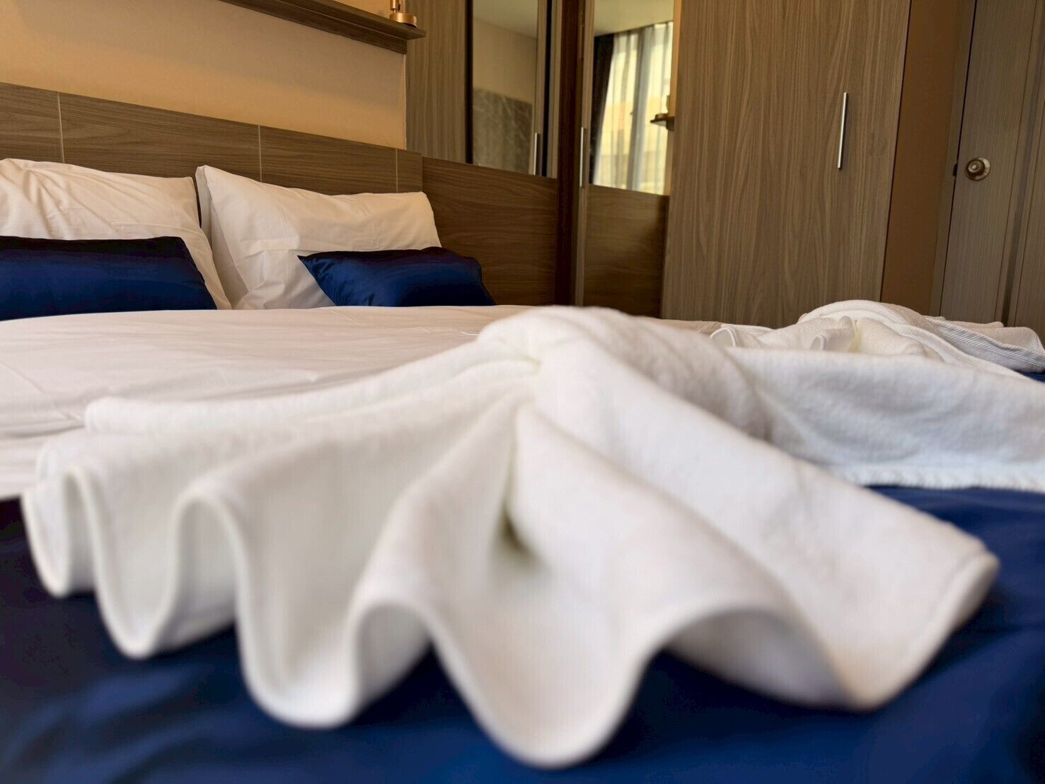 A neatly made bed with white bedding and blue pillows, a wooden headboard, and a wardrobe with a mirror.