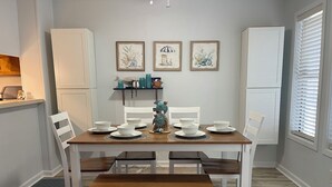 A dining area with a wooden table set for four, white chairs, and a bench. There are framed artworks on the wall, a small shelf with decorative items, and a window with blinds. - Pelican’s Perch: 3 minute walk to the beach, shops, restaurants and pavilion. (North Myrtle Beach)