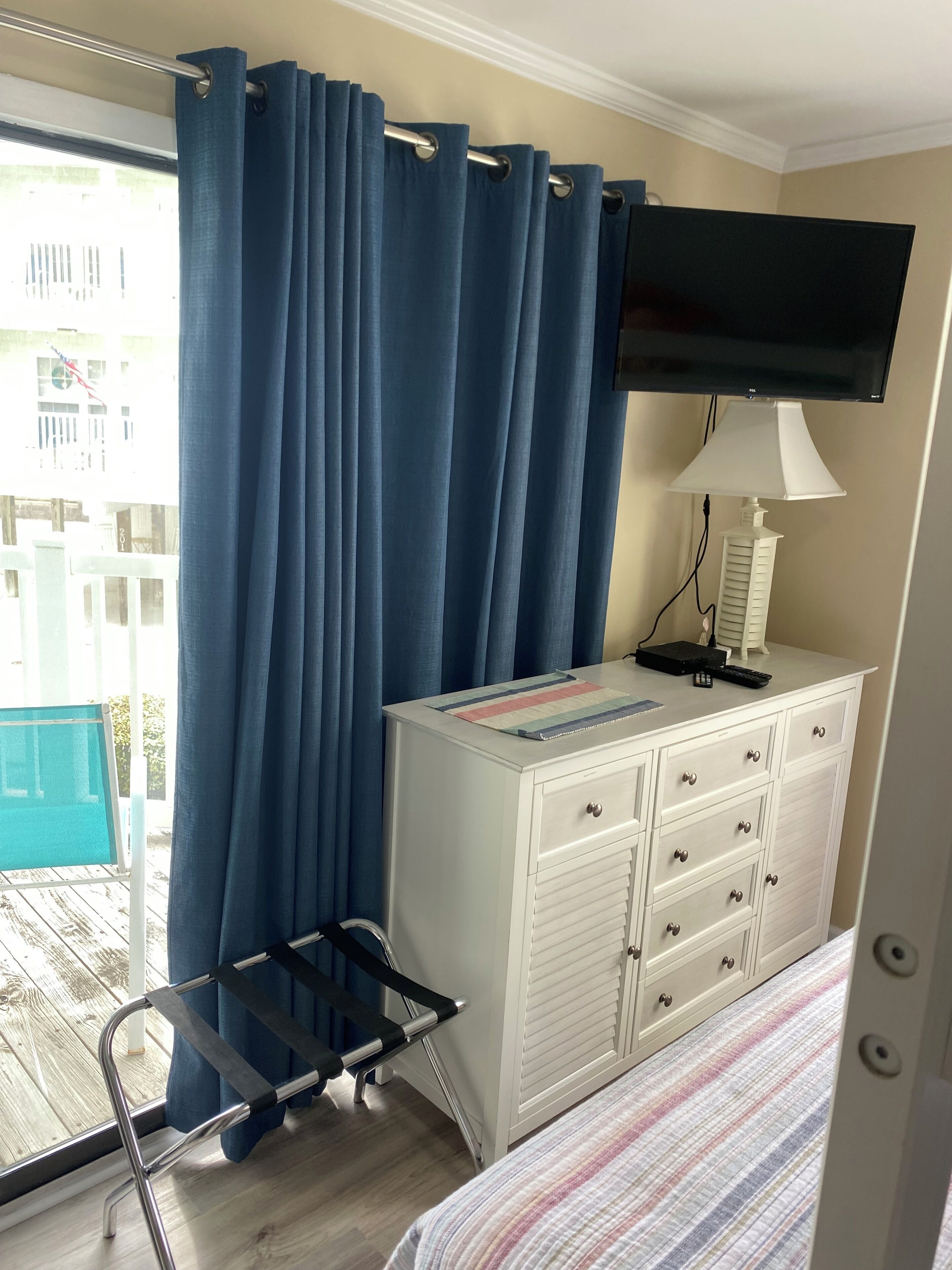 A hotel room with a bed, a white dresser, a TV mounted on the wall, and a balcony with blue curtains.