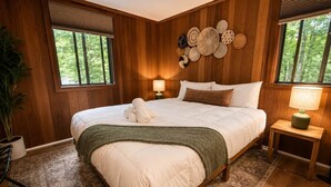 A wooden-paneled bedroom with a large bed, two bedside lamps, a wall of woven baskets, and a view of trees through the windows. - Pocono Basecamp | Hot Tub Chalet + Big Game Room (Tunkhannock Township)