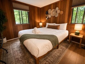 Ein schlächlich eingerichtetes Schlafzimmer mit einem großen Bett, zwei Nachttischlampen, einer Wand voller gewebter Körbe und Blick auf Bäume durch die Fenster.