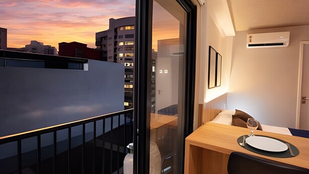 A modern room with a balcony, a dining area with a table and chairs, and a view of the cityscape at sunset. - Studio Elegance Next Pamplona (São Paulo)