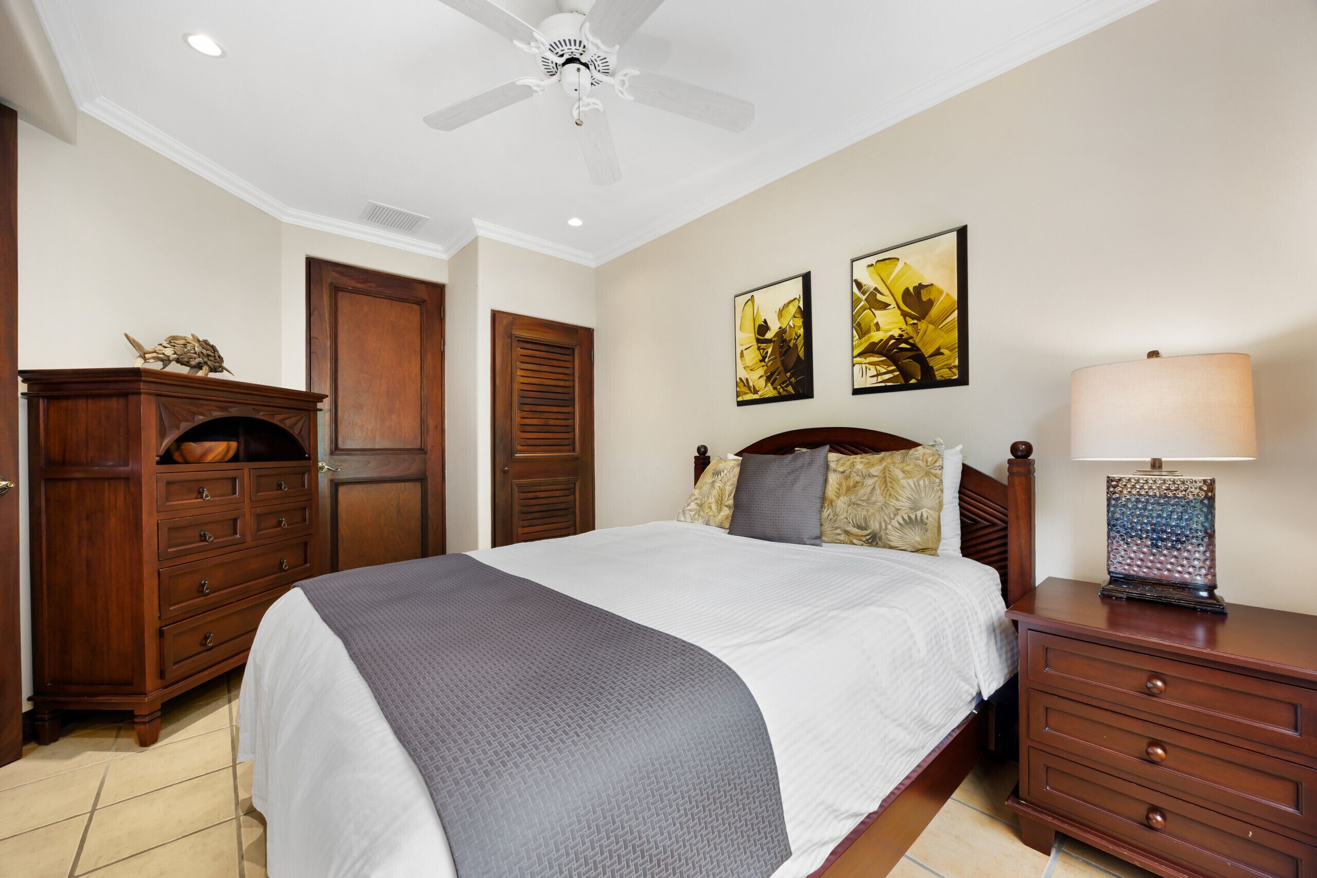 Une chambre à coucher comprenant un lit, une commode, une table de chevet, un ventilateur de plafond et des œuvres d’art accrochées au mur.