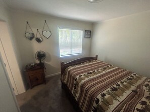 Ein Schlafzimmer mit Bett, Nachttisch, Ventilatoren, einem Fenster mit Jalousien und Wanddekorationen, darunter herzförmige Elemente.