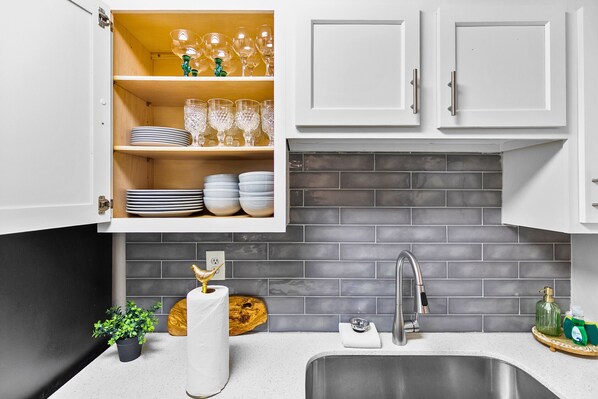A kitchen with white cabinets, a grey tiled backsplash, a stainless steel sink, and a shelf with glassware and plates.