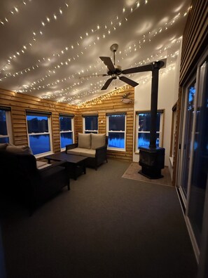 A cozy room with a ceiling fan, string lights, a fireplace, and large windows. - 3-bedroom lake house minutes from Pictured Rocks boat cruises and Munising! (Wetmore)