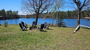 A lakeside outdoor seating area with Adirondack chairs and a small table, surrounded by trees and a clear view of the lake. - 3-bedroom lake house minutes from Pictured Rocks boat cruises and Munising! (Wetmore)