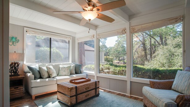 A room with a ceiling fan, a sofa, a wicker coffee table, and large windows offering a view of greenery. - Relax & Unwind|2BR Sea Pines Getaway (Hilton Head Island)