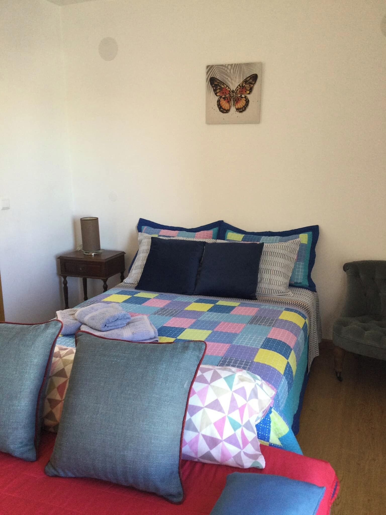 Una camera da letto con un letto, cuscini, un comodino, una sedia e un quadro raffigurante una farfalla appesa al muro.