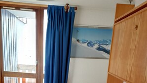 Ett hotellrum med en säng, en sänggavel i trä, en målning som föreställer ett snöigt berg och en balkong med blå gardiner.