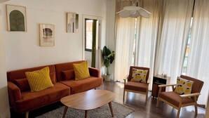 Ein Wohnzimmer mit einer Couch, Sesseln, einem Couchtisch und einem großen Fenster mit Vorhängen.