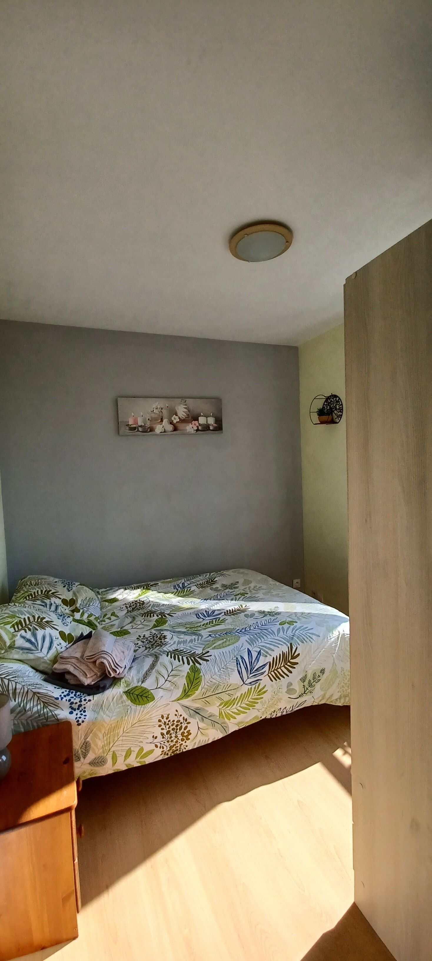A room with a bed, a small wooden dresser, a wall-mounted picture, and a ceiling light fixture.