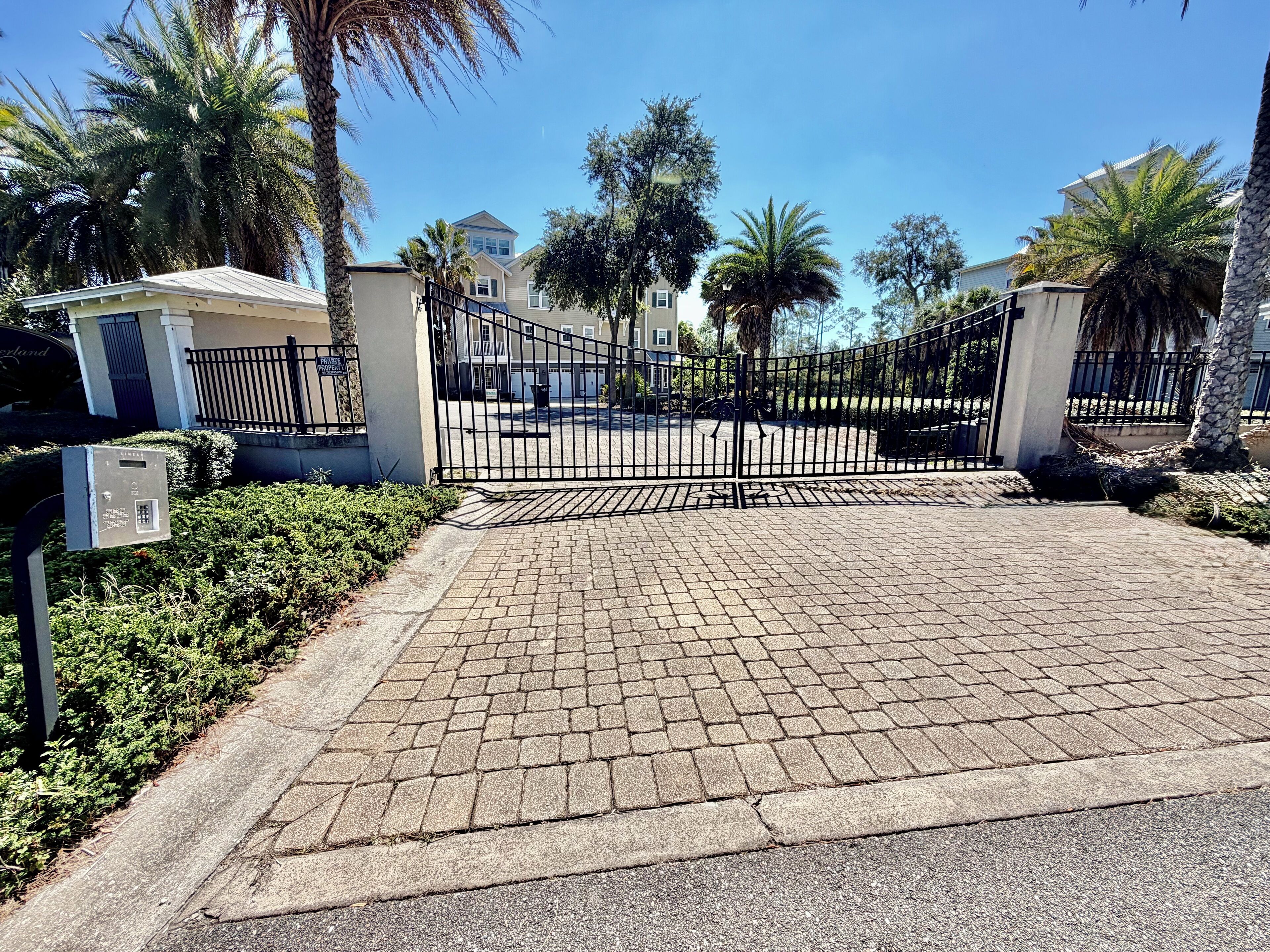 A gated entrance with a paved driveway leading to a building, surrounded by palm trees and a clear sky.