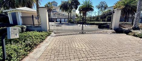 A gated entrance with a paved driveway leading to a building, surrounded by palm trees and a clear sky.