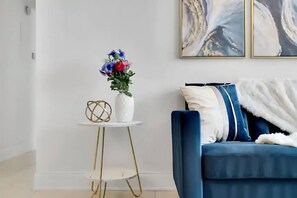Ein modernes Wohnzimmer mit einem blauen Sofa, einer weißen Vase mit Blumen und einem kleinen weißen Tisch mit goldenen Beinen.