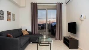 A modern living room with a sofa, a glass coffee table, a TV, and a view of the cityscape through large windows. - Pilotlux Apartments (Chania)
