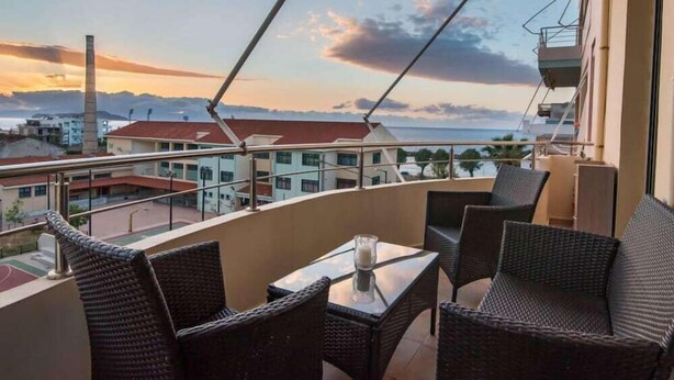 A balcony with wicker furniture and a glass table, overlooking a cityscape with buildings and a distant view of the sea. - Pilotlux Apartments (Chania)