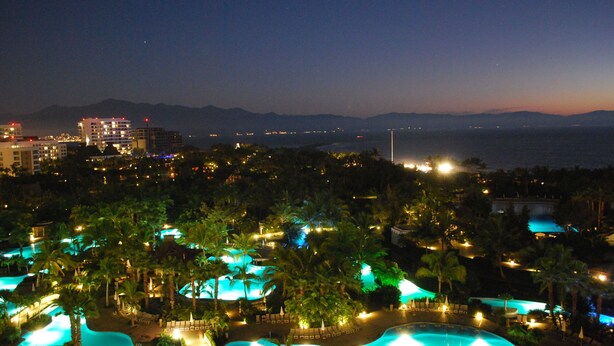 Un resort con piscina, palmeras y vistas al océano y montañas de noche.